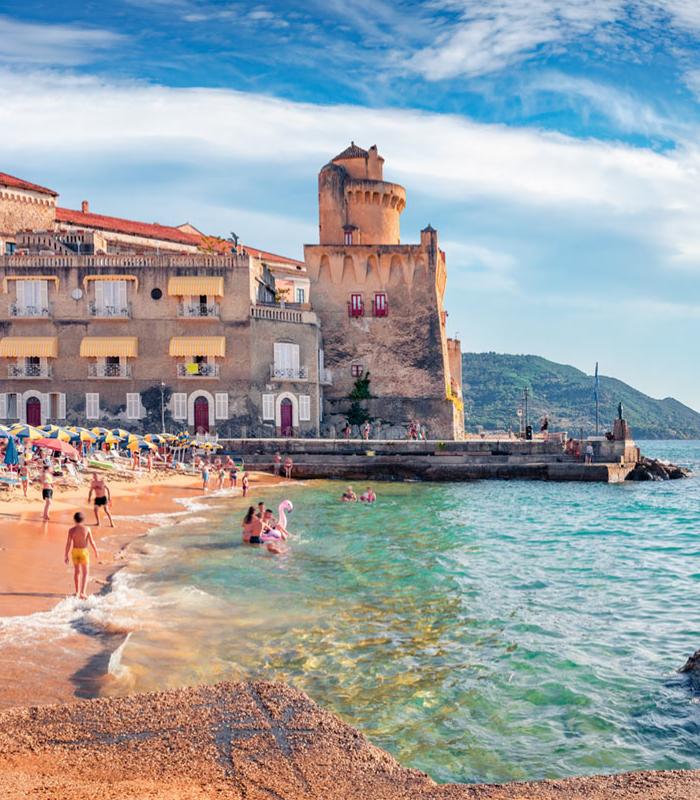 Spiaggia affollata con mare cristallino e antica torre in un borgo pittoresco.